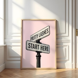 May include: Framed print with two street signs saying "Pretty Lashes" and "Start Here" against a pink backdrop. The signs are on a black post. The frame is light wood-coloured.