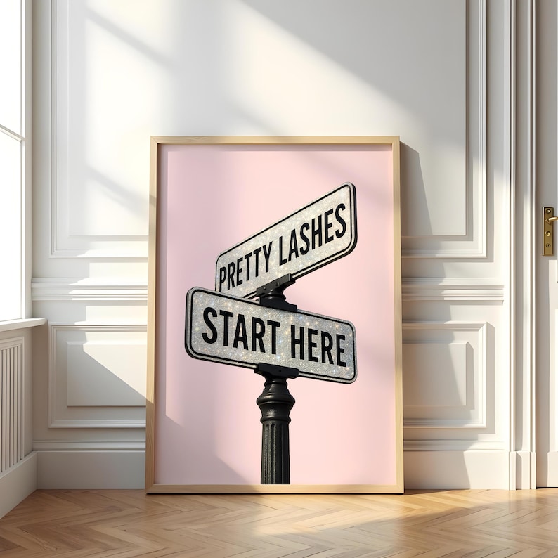 May include: Framed print featuring two street signs that read "Pretty Lashes" and "Start Here" against a pink background. The signs are mounted on a black post. The frame is a light wood color.