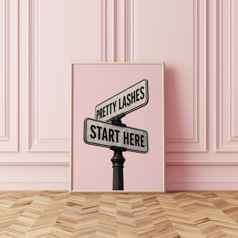 May include: A framed print featuring two street signs with the phrases "Pretty Lashes" and "Start Here" in black text. The signs are mounted on a black post against a pink background.