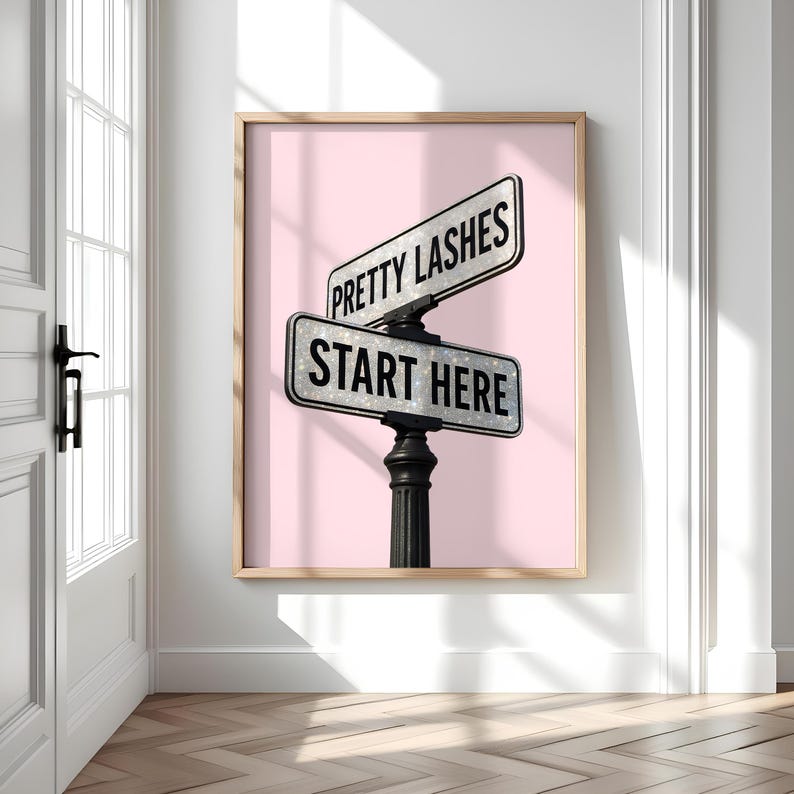 May include: A framed print featuring a pink background with a street sign that reads "PRETTY LASHES" and "START HERE" in black text. The sign is silver and mounted on a black post. The frame is a light brown color.