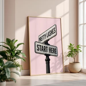 May include: A framed print featuring a street sign design with the text "Pretty Lashes" and "Start Here" on a pink background. The sign is black and white with a glittery effect, mounted on a black post. The frame is gold.