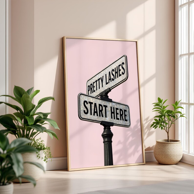 May include: A framed print featuring a street sign design with the text "Pretty Lashes" and "Start Here" on a pink background. The sign is black and white with a glittery effect, mounted on a black post. The frame is gold.
