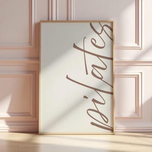 May include: A framed print with the word "pilates" in a stylish, cursive font. The text is a warm brown color against a cream-colored background. The frame is a light wood tone. The print is leaning against a light pink wall.