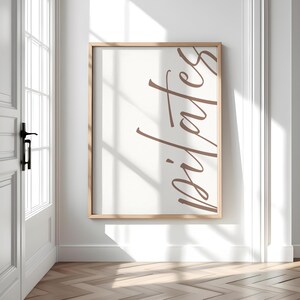 May include: A framed print with the word "pilates" in a stylish, cursive font. The text is in a warm brown color against a cream-colored background. The frame is a light wood tone, and the print is displayed on a white wall.