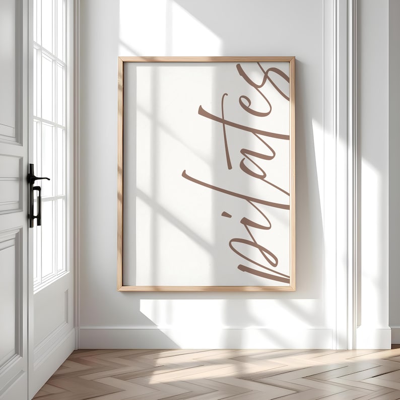 May include: A framed print with the word "pilates" in a stylish, cursive font. The text is in a warm brown color against a cream-colored background. The frame is a light wood tone, and the print is displayed on a white wall.