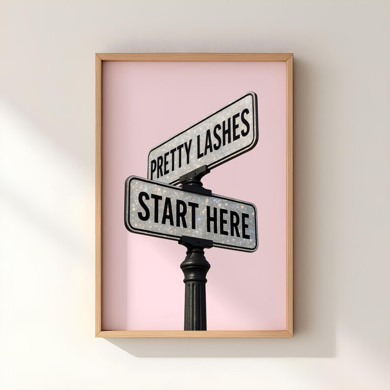 May include: A framed print featuring two street signs with the phrases "PRETTY LASHES" and "START HERE" in black text. The signs have a glittery texture and are set against a pink background. The frame is a light brown color.