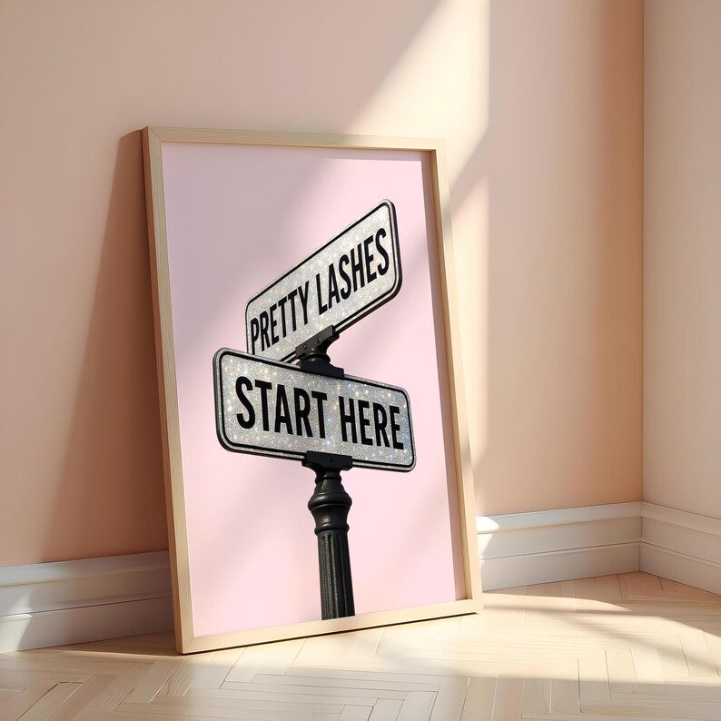 May include: Framed print featuring a pink background with a street sign that reads "PRETTY LASHES" and "START HERE" in black text. The sign is mounted on a black post. The frame is light brown.
