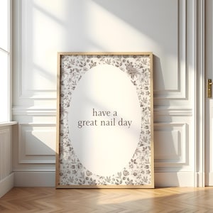 May include: A framed print with the text "have a great nail day" in a light brown font. The print has a floral pattern border and is set against a white background. The frame is a light wood color.