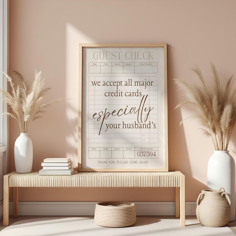 May include: A framed guest check print with the text "we accept all major credit cards, especially your husband's." The print is displayed on a woven bench with decorative vases and a small basket.