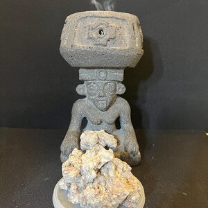 May include: A gray stone incense burner in the shape of a seated figure with a square top. Smoke rises from the top. A pile of light brown incense sits on a clear plastic lid in front of the figure.