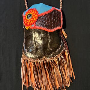 May include: A small, brown leather bag with a beaded sunflower design and a fringe trim. The bag is made from alligator skin and has a blue leather flap.