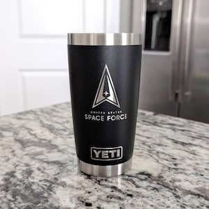 May include: Black insulated tumbler with a silver rim and base. The tumbler features the United States Space Force logo and text. The brand name "YETI" is displayed below the text. The tumbler is cylindrical and sits on a speckled countertop.