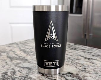May include: Black insulated tumbler with a silver rim and base. The tumbler features the United States Space Force logo and text. The brand name "YETI" is displayed below the text. The tumbler is cylindrical and sits on a speckled countertop.
