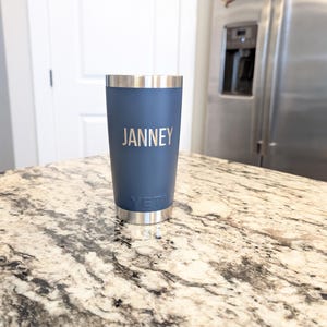 May include: A navy blue insulated tumbler with the name "JANNEY" printed in silver. The tumbler has a silver rim and base, and the brand name "YETI" is visible near the base. The tumbler is sitting on a granite countertop.