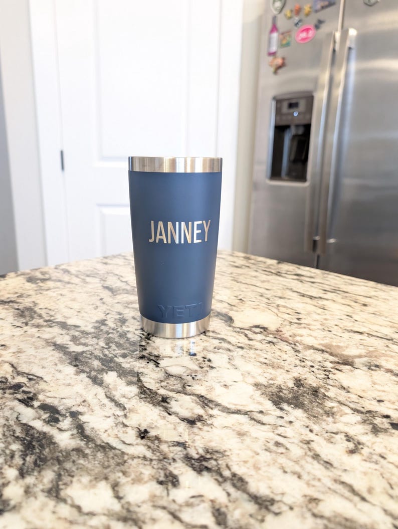 May include: A navy blue insulated tumbler with the name "JANNEY" printed in silver. The tumbler has a silver rim and base, and the brand name "YETI" is visible near the base. The tumbler is sitting on a granite countertop.