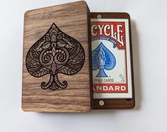 May include: A wooden playing card box with an open lid, revealing a deck of Bicycle playing cards. The lid features an intricately carved spade design. The Bicycle card box is made of dark wood and has a magnetic closure.