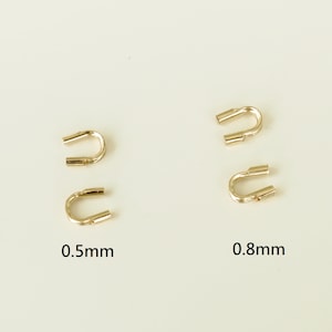 May include: Four gold-coloured U-shaped jewellery findings. Two are labelled 0.5mm and two are labelled 0.8mm. The findings have a U-shape with open ends. The background is a solid, light beige colour.