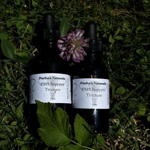 May include: Two brown glass bottles with dropper tops, labeled "Pacha's Naturals PMS Support Tincture". The bottles are sitting on a bed of green grass.