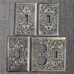 May include: Four decorative outlet covers with an ornate, silver-coloured design. The covers feature a floral pattern and are designed to fit standard electrical outlets.