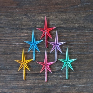 Peut inclure: Sept étoiles de Noël colorées en forme d'étoile dans différentes couleurs, dont le rouge, le bleu, le violet, le jaune et le vert, sur un fond de bois foncé.
