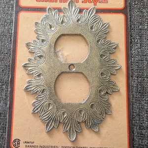 May include: A metallic gold-toned electrical outlet wall plate with an ornate, leaf-like embossed border. The plate is mounted on a rectangular cardboard backing labelled Charm-n-Style, Banner Industries, and Danbel Industries Inc.