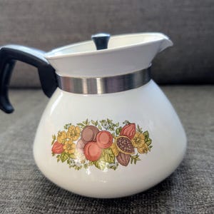 May include: White ceramic coffee pot with a black handle and lid. The pot features a decorative band of silver metal and a floral design with brown, orange, and yellow accents. The pot is designed for brewing coffee.