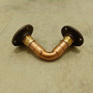 Pode incluir: Um suporte de prateleira de tubo de cobre com flanges de ferro preto. O tubo é dobrado em um ângulo de 90 graus e tem um acessório de latão em cada extremidade.