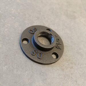 May include: Black metal pipe flange with a threaded opening and three holes. The flange is labeled "DS" and "OS".