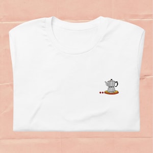 Könnte beinhalten: Weißes T-Shirt mit einem gestickten Design einer silbernen Espressokanne und zwei roten Punkten. Die Espressokanne steht auf einem gelben Sockel. Das T-Shirt ist auf einer rosa Oberfläche gefaltet.
