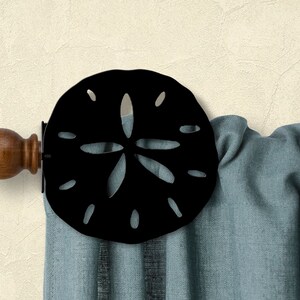 May include: Black metal curtain tieback with a sand dollar design. The tieback is attached to a wooden curtain rod with a brown finial.