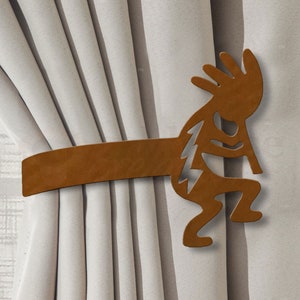 May include: A brown metal curtain tieback shaped like a Native American dancer holding a flute. The tieback is holding back a set of white curtains.