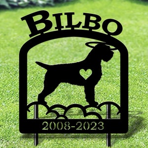 May include: Black metal garden memorial with a silhouette of a dog, a halo, and a heart. The memorial has the name "Bilbo" and the dates "2008-2023" inscribed on it.
