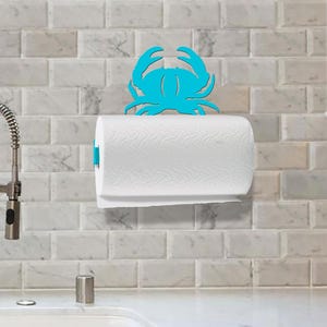 May include: A wall-mounted paper towel holder featuring a bright turquoise crab silhouette positioned above a standard roll of white paper towels with a textured pattern, mounted on a light-colored rectangular tiled kitchen backsplash.