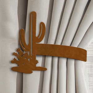 May include: Brown metal cactus curtain tieback. The tieback is shaped like a cactus with a long stem and multiple arms. The cactus is attached to a brown leather strap.
