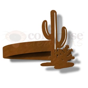May include: Rustic brown metal cactus napkin ring. The ring is shaped like a tall cactus with smaller cacti at the base.