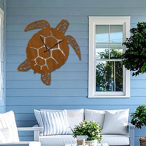 May include: A wall clock shaped like a sea turtle with a rust-colored metal finish and a cutout shell pattern. The clock features thin black hour and minute hands positioned at the center of the shell, mounted on a light blue horizontal siding wall.