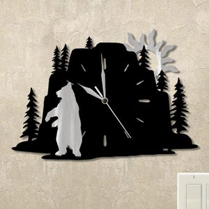 May include: A black and silver metal wall clock featuring a bear standing in front of a mountain silhouette. The clock face is black with silver hands and hour markers. Trees and a sun-like design are also included.