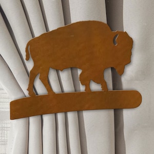May include: A brown metal buffalo silhouette curtain tieback. The buffalo is standing on a brown metal base.