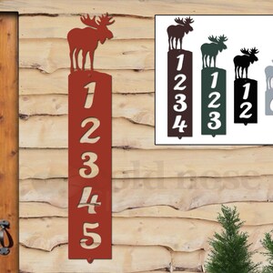 May include: A red metal address sign with the numbers 1, 2, 3, 4, and 5 cut out. A silhouette of a moose is at the top. The sign is mounted on a wooden wall. Additional signs with different numbers and colors are also shown.