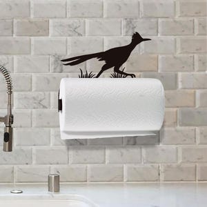 May include: A black metal paper towel holder featuring a silhouette of a roadrunner bird standing in grass. The holder is mounted on a wall and supports a roll of white paper towels with a textured pattern on the surface of the paper.
