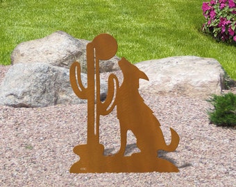 May include: Metal silhouette of a coyote howling at a cactus, both are rusty brown in color. The sculpture is on a bed of gravel with green grass and rocks in the background.