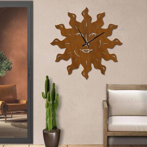 May include: A sun-shaped wall clock with a rust-colored metal finish, featuring a stylized face with cut-out eyes and a mouth. The clock has thin black hour, minute, and second hands positioned at the center of the sun's face, creating a decorative piece.