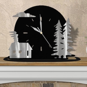 May include: A black and silver metal clock with a cabin, trees, and clouds cut out. The clock face is black with silver hands and a silver rim. The clock is on a black base.