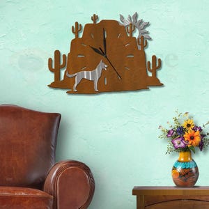 May include: A wall clock featuring a brown desert landscape silhouette with cacti and a silver sun. A silver dog stands in the foreground. The clock has black hands and horizontal cutouts for hour markers, creating a desert-themed wall decor piece.