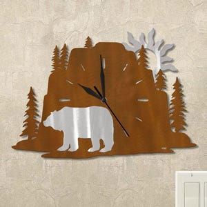 May include: A metal wall clock featuring a bear silhouette in front of a mountain scene with trees and a sun. The clock has a brown finish with a silver bear and sun. The clock hands are black.