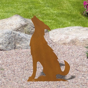 May include: A rusty brown metal silhouette of a howling wolf, positioned on a bed of small pebbles. The wolf is in a seated position with its head raised, set against a backdrop of green grass, rocks, and plants. The metal art piece is designed for outdoor use.