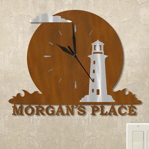 May include: Metal wall clock featuring a lighthouse design. The clock has a brown circular background with a silver lighthouse and cloud. The clock hands are black, and the text "MORGAN'S PLACE" is at the bottom.