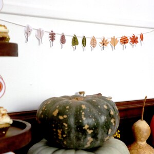 May include: An autumnal display with a garland of leaf cutouts in various colours, cupcakes with frosting, and pumpkins. The garland is strung across a white wall, and the pumpkins are in shades of green and orange.