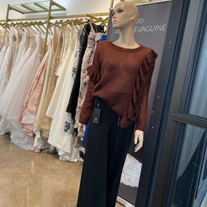 Puede incluir: Un maniqu&iacute; con un su&eacute;ter color &oacute;xido con cuello de volantes y pantalones negros de pierna ancha. El maniqu&iacute; est&aacute; de pie frente a un estante de vestidos y un letrero que dice "SE EVAGUINE".