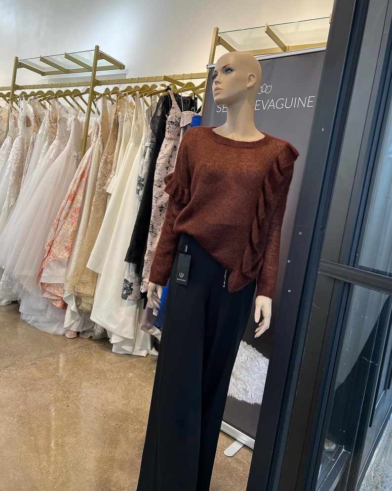 Puede incluir: Un maniqu&iacute; con un su&eacute;ter color &oacute;xido con cuello de volantes y pantalones negros de pierna ancha. El maniqu&iacute; est&aacute; de pie frente a un estante de vestidos y un letrero que dice "SE EVAGUINE".